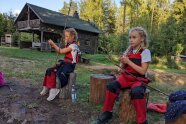 zwei Mädchen sitzen auf Holzhockern und legen Hefeteig für Stockbrot um ihre Grillstöcke