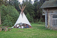 ein Tipi mit vielen sitzenden Menschen darin, daneben eine Stück einer Blockhütte