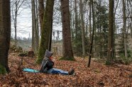 Person mit Winterkleidung und Mütze sitze auf einer Isomatte mitten im Wald und trinkt Tee